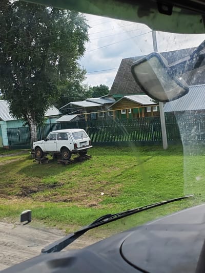 A white Lada Niva is elevated on wooden blocks on a grassy area next to a residential street in Novye Vyselki, Russia. The car is facing left. Behind the car is a green wooden fence, and behind that are several houses with corrugated metal roofs. A large tree is to the left of the car. The photo appears to be taken from inside another vehicle, with part of a windshield wiper and the dashboard visible in the foreground. The sky is cloudy.