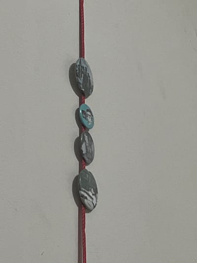 Four oval-shaped decorative charms, featuring black and white patterns and one with a teal background and a person's face, are suspended vertically on a thin red string against a plain gray wall. The charms are spaced out evenly along the string. The scene is indoors, with no visible indications of the city of San Luis, Peru.