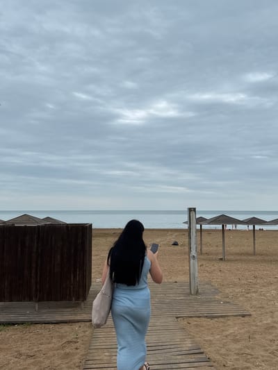 A woman with long black hair, wearing a light blue dress and carrying a beige tote bag, walks on a wooden boardwalk towards the sea on an overcast day at a beach in Derbent, Russia. She is holding a black smartphone in her right hand. In the background, there are several wooden beach umbrellas and a few people scattered on the sandy beach. The calm sea stretches to the horizon under a cloudy sky.