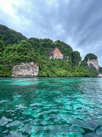 A wide shot captures the stunning seascape off the coast of Ko, Thailand, under a cloudy sky. Verdant, jungle-covered limestone cliffs dominate the background, with sections of exposed rock displaying striking orange and white striations. The foreground is filled with crystal-clear turquoise water, its surface rippled by gentle waves. The clarity of the water reveals a glimpse of the underwater world beneath. No people or man-made structures are visible in this naturalistic landscape.
