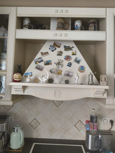 The image shows a kitchen shelf with various decorative items. On the top shelf, there are several mugs, some of which appear to be souvenir beer steins. The middle section features a triangular surface decorated with numerous refrigerator magnets, depicting landmarks, animals, and other designs. Below this, a wooden shelf displays a collection of small figurines, including an angel, a cat, and a Daruma doll, along with a salt grinder and other decorative objects. The background consists of tiled walls and cabinetry. The scene is set in a kitchen, and based on the context of the location, this kitchen is likely located in Мещерино, Russia.