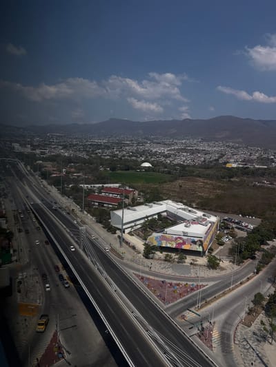This aerial-perspective image depicts a segment of Tuxtla Gutiérrez, Chiapas, Mexico, dominated by a modern urban landscape and distant mountains under a partly cloudy sky.
The central feature is a multi-lane elevated highway, likely part of the Libramiento Norte Poniente, which traverses the scene diagonally from the lower left to the middle right. The highway is dark-paved with clear white lane markings and is supported by visible white pylons and cable stays. Numerous vehicles, predominantly cars and a few trucks, are in transit on both directions of the highway. Beneath and adjacent to the elevated structure, a complex network of ground-level roads and intersections facilitates local traffic. A yellow utility truck is visible on a lower road segment near the bottom left.
To the right of the elevated highway lies a prominent, angular, multi-story building identified as the Museo de Ciencia y Tecnología de Chiapas (MUCH). The building features a predominantly white facade and roof, with multiple HVAC units on its surface. A significant portion of its exterior wall displays a large, colorful mural depicting stylized human figures and abstract forms in vibrant hues. Adjacent to the building, a large parking lot contains numerous parked automobiles.
Further into the mid-ground, a dense urban sprawl of residential and commercial structures extends towards the mountain range in the distance. A distinct white, dome-shaped building, likely the Planetario de Tuxtla Gutiérrez, is visible within a green field area in the middle distance. Several other buildings with red-tiled roofs are situated behind the museum.
The background is defined by a range of dark, forested mountains. The sky overhead is bright blue with scattered white cumulus clouds, indicating daytime conditions. Small, indistinct human figures are discernible on sidewalks near the road intersections and the museum building. The overall scene suggests a vibrant, active urban environment with ongoing development and infrastructure.
