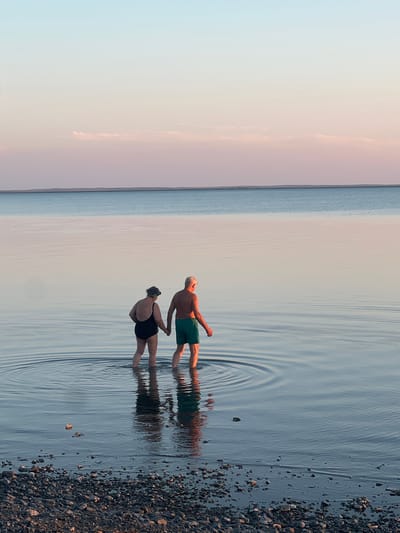 An elderly couple walks hand-in-hand into the shallow waters of the Caspian Sea near Karashygan, Kazakhstan, at sunset. The sky transitions from a pale blue at the top to soft pink and lavender hues near the horizon. The water is calm, with gentle ripples emanating from the couple's movement. The foreground consists of a rocky shoreline. The man wears green swim shorts, and the woman wears a black swimsuit and goggles on her head. Their reflections are visible on the water's surface.