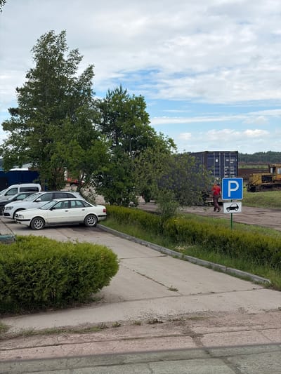 A paved parking lot is visible with several cars. In the foreground, a dense green shrub borders the lot. Further back, a white sedan is parked alongside a white SUV. To the right of the cars, a blue parking sign with a white "P" and a car icon is displayed on a post. A man in a red shirt and brown pants is walking away from a dark shipping container, which is positioned behind the parking sign. A yellow bulldozer is visible in the background to the right. The environment appears to be an outdoor industrial or logistical area with trees and a cloudy sky.