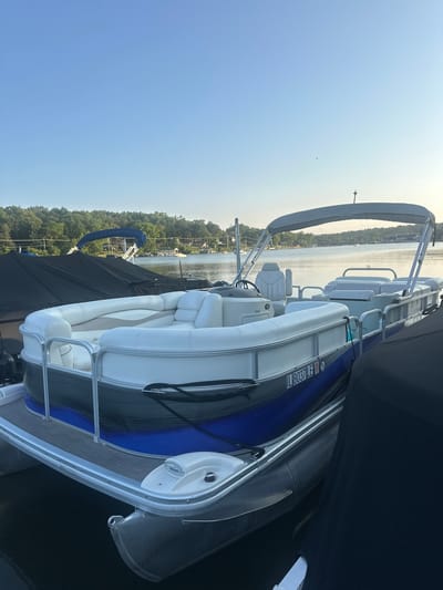 A pontoon boat, identified by the registration "IL 8037 LE", is docked on a body of water, likely a lake or river, in Algonquin, Illinois, USA. The boat features white upholstery and a blue and gray exterior. It is positioned alongside other boats, some of which are covered with black tarps. The background shows a tree-lined shore with houses and a clear blue sky. The scene appears to be set during daytime, with the sun casting a gentle light.