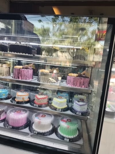 The image shows a refrigerated display case filled with various cakes, likely in a bakery. The display case is divided into multiple shelves. On the top shelf, there are several dark chocolate cakes with white frosting and cherry toppings. On the second shelf, there are several decorative cakes with dripping icing and "Happy Birthday" toppers. These cakes are various colors including pink, white, and beige. On the third shelf, there are smaller, individual-sized cakes. These include a blue cake with yellow stars, an orange and white cake, a pink and white cake, a yellow and white cake, and a purple and white cake. On the bottom shelf, there are more round cakes, including a pink cake, a white cake, a green cake, and a dark cake with silver sprinkles visible on the far right. The cakes are predominantly round and covered in plastic domes for protection. The reflection in the glass of the display case shows trees and possibly a building with a sign that says "ITO". The overall environment appears to be a commercial establishment. Given the context of Krian, Indonesia, this is likely a bakery in that city.
