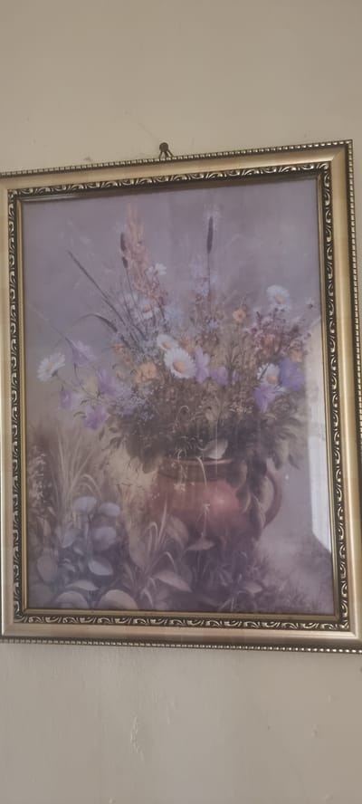 This media file features a framed painting of a floral arrangement. The painting depicts a bouquet of flowers, primarily white and purple daisies, along with other smaller blooms and greenery. The flowers appear to be arranged in a rustic setting, possibly in a jug or vase that is partially visible in the background, rendered in warm earthy tones. The background of the painting is a soft, muted purple, giving the impression of a gentle, atmospheric setting. The painting is housed in an ornate, gold-toned frame with a patterned border. A small, dark circular object, possibly a nail or hook, is visible on the wall to the left of the frame, indicating it is hung. The overall mood of the painting is serene and evokes a sense of natural beauty. No people are present in the artwork. The setting appears to be indoors, as it is a framed artwork on a wall. There is no visible text in the image.