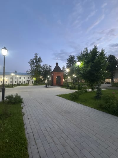 This is a nighttime exterior shot of a paved courtyard area in the city of Vereya, Russia. In the foreground, a wide, gray brick pathway leads into the scene, bordered by a strip of green grass. Several ornate street lamps with glowing bulbs line the pathway.
To the left of the pathway, a long, white building with numerous windows and a blue roof is visible. In the center of the courtyard, a small, red brick chapel or shrine with a dark, domed roof and a cross on top stands prominently.
Behind the chapel, a variety of green trees create a natural backdrop. The sky above is a deep twilight blue with wispy clouds. The scene appears serene and well-maintained, suggesting a public or religious space. There are no people or animals visible in the image.