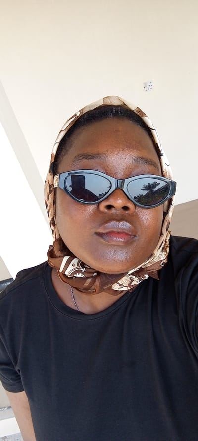 An individual with dark skin, appearing to be an adult female, is depicted in a close-up, chest-up portrait shot in Ijotan, Nigeria. She faces directly forward, wearing dark, oval-shaped sunglasses that obscure her eyes. Her lips are full with a reddish tint, and her skin exhibits a natural sheen. She is dressed in a plain black crew-neck t-shirt. A patterned headscarf, featuring shades of beige, brown, and cream with an elaborate design, covers her hair and is tied loosely around her neck. A thin, silver-colored chain is visible around her neck, beneath the scarf.
The background primarily consists of a light beige or off-white wall. To the far left, a bright white vertical architectural element is visible. In the upper right quadrant of the background, a white multi-standard electrical socket is mounted on the wall. Reflections in the sunglasses show a blurred silhouette of a person holding a device, along with the distinct outlines of buildings and lush green foliage, including what appear to be palm trees, under a bright sky. The scene is illuminated by daylight, suggesting an outdoor or semi-outdoor location within the city of Ijotan.