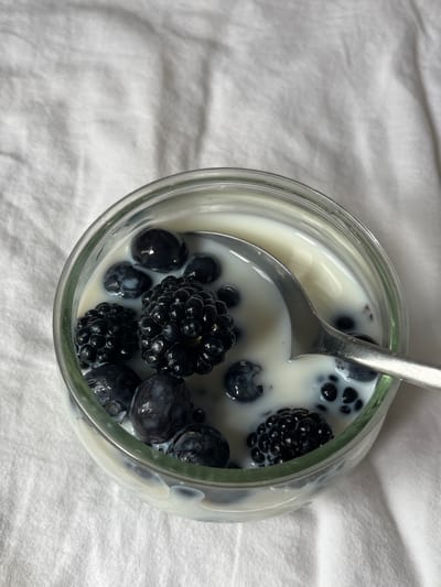 The image presents a high-angle, close-up view of a glass jar or cup containing a serving of milk or yogurt with fresh berries. The main subjects are the food items: a generous quantity of dark blueberries and blackberries are visible, submerged in a creamy white liquid, likely milk, yogurt, or a similar dairy product. A silver spoon is partially submerged within the jar, positioned towards the top left, suggesting the dish is being prepared for consumption or is already being eaten.
The berries appear plump and fresh, with the blackberries showing their characteristic segmented structure. The liquid has a smooth texture, gently coating the fruit. The glass container is simple and clear, allowing full visibility of its contents.
The setting is understated, with the jar resting on a soft, light-colored, possibly white or off-white, fabric surface that has subtle wrinkles and folds, creating a textured background. This background occupies the majority of the left side and extends around the jar, suggesting an indoor, casual environment like a bed or a cozy table setting.
There is no specific activity explicitly shown beyond the presence of the prepared food, implying a quiet moment for a meal or snack. The time of day is not overtly indicated but the type of food suggests breakfast or a light refreshment. There are no people visible, and therefore no emotions are directly conveyed. No text is visible in the image. While the location context provided is Aarhus, Denmark, there are no visual cues within the image itself to confirm this geographical location.