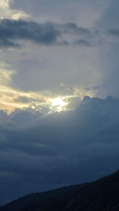 The image shows a cloudy sky with the sun shining through a gap in the clouds. A dark, possibly forested, hill or mountain is visible on the right edge of the frame. The location is Ba Ngoi Ward, Vietnam. There are no people or immediate actions visible in the frame. The primary subjects are the sky, clouds, sunlight, and a landform. The environment appears to be outdoor and possibly rural, given the presence of the hill/mountain.