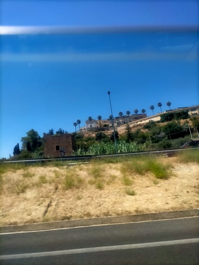The image depicts a view of a rural hillside with buildings and vegetation, taken from a moving vehicle. In the foreground, there is a paved road and a dry, grassy embankment. The middle ground features a more lush hillside covered in green foliage and trees, with a prominent dilapidated stone structure resembling a tower or ruin on the left side. In the background, on top of the hill, a row of buildings with palm trees in front of them is visible. The sky is clear and blue. The scene is captured on a sunny day. The overall impression is a landscape view of the outskirts of Sperone, Italy.