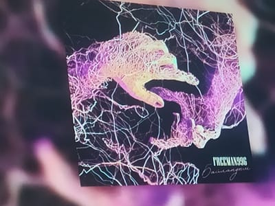 This image shows a close-up of a digital artwork, likely album art, with an abstract, organic design. The artwork features what appears to be a stylized human hand interwoven with a network of delicate, white, and pinkish-purple tendrils or fibers against a dark background. The colors are vibrant and have an almost bioluminescent quality. In the bottom right corner of the artwork, two lines of text are visible. The top line is in a bold, sans-serif font and reads "FREEMAN996". Below this, in a cursive script, is another line of text that is partially obscured but appears to say "Dainenugnu". The artwork is displayed on a screen, indicated by the slight blur and the way it is angled within the frame, suggesting it is being viewed on a device. The surrounding environment is out of focus and consists of dark, blurred shapes with subtle purple and greenish hues, likely representing the ambient lighting or background of the room where the screen is located. There is no identifiable information within the image that connects it to Otuzuul, Kyrgyzstan, or any specific urban environment.