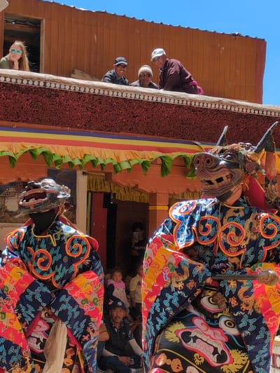 The image captures a scene during a cultural event, possibly a festival or performance, in Pibiting, India. In the foreground, two individuals are dressed in elaborate, traditional costumes and masks. The person on the right wears a mask resembling a fierce animal with horns and a snout, paired with a vibrant robe adorned with swirling patterns and dragon motifs in blues, reds, yellows, and oranges. The person on the left is also masked, wearing a darker, open-mouthed mask and a similarly patterned costume. Both figures appear to be performing or posing.
In the middle ground, a colorful banner with yellow, orange, red, and green stripes hangs across the facade of a building. Below the banner, more people are visible, some seated and watching the performance. A child in a pink and white outfit is standing, looking towards the foreground figures. Another man, wearing a dark baseball cap, is seated in the lower right portion of the frame, looking towards the masked performers.
In the background, a woman wearing sunglasses and a light-colored shirt is leaning on a railing on an upper level of a building. She appears relaxed, observing the scene below. Other individuals are also visible on this upper level, some looking down at the activity. The architecture features wooden structures and reddish-brown textured walls, suggesting a traditional Indian style. The sky above is clear and blue. The overall atmosphere appears to be festive and communal, with a blend of performers, spectators, and the distinct cultural attire and architecture of Pibiting.