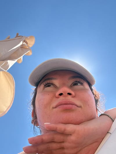 A low-angle, close-up shot captures a woman wearing a light-colored baseball cap and looking upwards against a clear blue sky. Sunlight beams from behind her head, creating a halo effect. Her chin rests on her hand, which is adorned with a beaded bracelet. To the left of the frame, a light-colored fabric, possibly part of a beach umbrella, is visible. The image appears to be taken outdoors on a sunny day, with no discernible urban or natural landmarks to indicate a specific location within Derbent, Russia.
