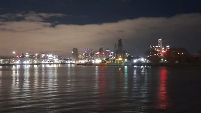 The media file shows a blurry, night-time, cityscape reflected on a dark body of water. The cityscape is comprised of numerous buildings of varying heights, illuminated by many lights. Some buildings appear to be skyscrapers. There are also what appear to be ships or boats docked along the waterfront, with lights visible on them as well.
The time of day is clearly night, as evidenced by the dark sky and the illuminated buildings. The sky is partly cloudy, with some patches of light illuminating the clouds. The water is choppy, with ripples and waves disturbing the reflections of the city lights. The reflections are elongated and distorted due to the movement of the water.
The overall mood of the image is somewhat atmospheric and possibly a bit melancholic, due to the darkness and the blurriness of the scene. It evokes a sense of urban nightlife, but the lack of sharp details makes it difficult to discern specific activities or emotions.
There is no visible text in the image. The location context of Dock Sud, Argentina, suggests an industrial or port area within a city. However, without clearer details, it's impossible to identify specific landmarks or confirm the precise location within Dock Sud. The image captures a general impression of a waterfront city at night.
