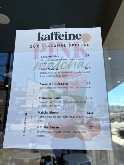 Here is a forensic description of the image.

The image shows a sign advertising seasonal specials at a café named "Kaffeine." The sign is taped to a window, likely of the café itself. The sign is white with black and pink text. The specials listed are:
- Coconut Kiss ($7)
- PINK Matcha ($8.5)
- Caramel Brûlée Latte ($7)
- Pistachio Bloom ($8.5)
- Matcha Bloom ($8.5)
- Matcha Breeze ($7)

The lower part of the sign reads "SIGNATURE DRINK." The text "BOLD, CREAMY & TOTALLY OBSESSED" is written in a semi-circular fashion on the left side of the sign. A graphic of a strawberry is visible at the bottom right of the sign.

The sign includes an endorsement indicating something or someone is 'the Goat!'

Based on publicly available information, there is a cafe named Kaffeine located in Redondo Beach, California, United States. The sign likely advertises specials at the Kaffeine located in Redondo Beach, California.