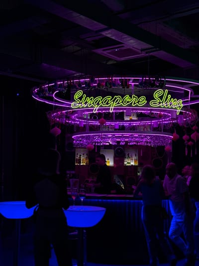 The image depicts the interior of a dimly lit bar with neon lighting. A prominent neon sign in the center reads "Singapore Sling" in yellow and purple. The bar itself is illuminated by blue lights from below and features bottles of alcohol displayed on shelves. Several people are visible, some standing at the bar, others at tables. The overall atmosphere is nocturnal and social. This scene is located at the Singapore Sling bar in Moscow, Russia.
