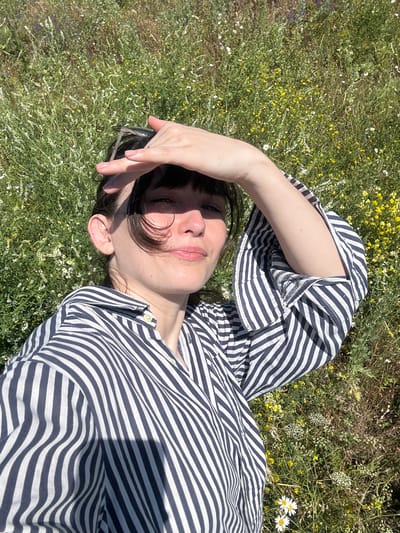 A woman is lying in a field of wild flowers, her head to the right of the frame. She is wearing a blue and white striped shirt and has her right hand shielding her eyes from the sun. Her left arm is bent and raised, with her hand resting on her forehead. The field is green and filled with small yellow and white flowers. The image was taken outdoors in Krasnyi Yar, Russia.