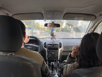 The image is taken from the interior of a car, looking out the front windshield. The driver, a man with short dark hair, is holding the steering wheel and looking at the road. A woman with long, dark hair is seated in the passenger seat, looking out the side window. She is wearing a military uniform, as is the driver. There is a rearview mirror on the windshield, showing a man with a short, dark beard driving another car, and a fuzzy, fluffy decorative object hanging from the rearview mirror.  The dashboard is visible and has several controls, including a radio, climate control, and other buttons and knobs. The car is parked in a parking lot, and there are other vehicles and pedestrians visible through the windshield. The location, based on the provided coordinates, is on a street near the intersection of Rager and Ha-Aliya Ha-Shniya Streets in Beersheba, Israel.