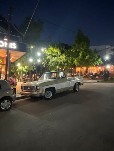 The image displays a nighttime street scene in City Bell, Argentina. A two-tone (light green/beige upper and white lower) Chevrolet C/K series pickup truck is parked on the asphalt pavement, facing left. To the left of the truck, the rear section of a grey compact car is partially visible. An establishment with an illuminated sign displaying "OMIS" is situated behind these vehicles, featuring outdoor seating beneath an awning. Multiple individuals are visible in this area, some seated at tables and others standing. One individual in a purple top and a face mask is seated at an outdoor table near the grey car. Further to the right, additional outdoor dining areas are visible, also populated by patrons and illuminated by string lights. Several trees with bright green foliage are present, illuminated by streetlights and ambient light. A bicycle is partially visible, parked on the right side of the street. Utility poles and overhead lines are visible against the dark sky.