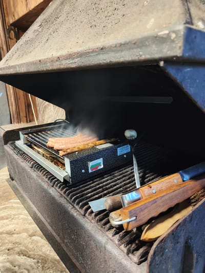 The image shows a close-up of a grill with several sausages cooking on it, producing visible smoke. The grill appears to be outdoors, possibly in a rustic setting given the wooden background and the weathered appearance of the grill itself. To the right of the sausages, a silver fork with a small white object on it is resting on the grill grates. There are also grilling tools with wooden handles piled on the side of the grill. A black metal hood is open above the grill. A small rectangular sticker on the front of the grill has a Bulgarian flag and text that reads "ПЛОЧАДОВОЕ БЪЛГАРИЯ" and "ПЛОЧАДОВОТО БЪЛГАРИЯ". The lighting suggests it could be daytime.