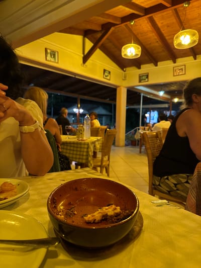 This image depicts a nighttime scene within a restaurant's covered dining area in Palaiokastritsa, Greece. In the immediate foreground, a table covered with a light-colored tablecloth displays a terracotta-colored ceramic bowl containing remnants of a consumed dish, likely moussaka, with ground meat and a white topping. To its left, a white plate holds a small, circular, light-brown food item, and another empty white plate with a fork is visible. A person's left arm, wearing a white shirt and a white wristband/watch with a ring on the hand, is partially visible at the left. Another person's right side, dressed in a black tank top and patterned pants, is visible on the right.

The midground features several tables with yellow and white checkered tablecloths and wicker chairs. Patrons are seated at these tables, blurred, engaged in dining. Objects such as water bottles, glasses, and cans are visible on the tables. The establishment has a wooden ceiling with exposed beams and recessed lighting. Overhead, multiple spherical glass light fixtures emit a warm, yellowish glow. Walls are light-colored, possibly cream, and adorned with framed pictures.

The background reveals the restaurant's open-air aspect, looking out into the dark night. Distant lights suggest other structures or street lighting, and vague outlines of foliage or buildings are discernible. The overall lighting is artificial and distinctly warm yellow, casting a predominant hue over the entire scene.