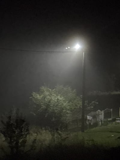 The image depicts a dimly lit, outdoor scene at night, possibly in the Neman Rustic Council area of Belarus. A single, bright streetlamp illuminates a misty or foggy atmosphere, casting a strong light to the left and obscuring details in the distance. In the foreground and midground, there are trees and vegetation, with a wooden fence visible in the background, along with what appears to be a small wooden structure. The overall impression is one of a rural or semi-rural setting enveloped in darkness and mist.