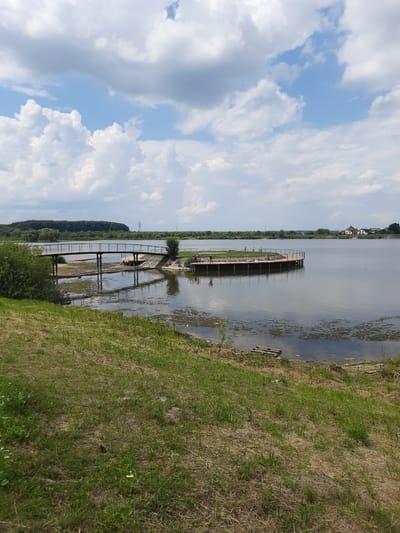 The image depicts a lakeside scene on a cloudy day. In the foreground, a grassy slope leads down to the water's edge. The lake occupies the middle ground, with several wooden structures extending into it. To the left, a pedestrian bridge crosses a shallow section of the lake or a connected body of water. Further to the right, a circular wooden platform with railings is situated in the lake, and several people are visible on it, some appearing to be sunbathing or relaxing. In the distance, a tree line borders the far side of the lake, with a few houses visible on the right bank. The sky is filled with a mix of white and grey clouds against a blue backdrop. The location is identified as the Terynivski Vody recreation area in Drohobych, Ukraine.