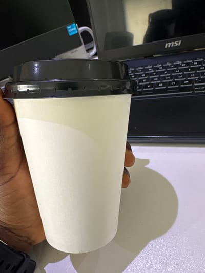 A close-up, slightly rotated shot captures a dark-skinned hand holding a plain white disposable cup with a black lid. The cup is held horizontally, positioned centrally in the frame. A black laptop, oriented vertically, is partially visible in the background, with its screen displaying the "MSI" logo prominently in white letters on its bezel. To the right of the laptop screen, part of the keyboard with visible keys is seen. Further to the left and slightly below the laptop, a device with a blue sticker, likely a brand emblem such as Intel, is visible. The hand holding the cup also wears a black smartwatch or fitness tracker on the wrist.
The setting appears to be indoors, likely at a desk or table, with the laptop and cup placed on a light-colored surface. A distinct, large shadow of the cup and hand is cast upon a light-colored wall or background surface to the right, suggesting the presence of a light source from the left. The overall lighting is indicative of an indoor environment. No specific activity beyond holding the cup is discernible, but it suggests the person might be about to drink. The scene is located in Gwaska, Nigeria.
