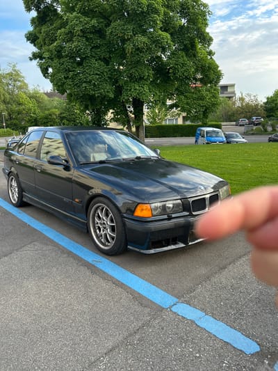 A black BMW 3 series is parked in a parking lot. The car is parked in a designated parking space marked by a blue line. There is a large tree in the background, along with a few buildings. The car is facing the camera, and the driver's side is visible. The car appears to be in good condition. There is a hand in the foreground, but the rest of the body is not visible. The image was taken at the coordinate location of latitude 47.544 and longitude 9.308 in the city of Amriswil, Switzerland.