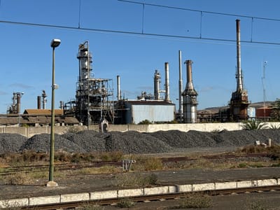 The image displays an industrial complex, identified as being in the city of Sidi Kacem, Morocco, under a clear, bright blue sky. In the foreground, multiple railway tracks run horizontally across dry earth interspersed with sparse vegetation, bordered by a white-painted concrete platform edge at the bottom. A tall, grey streetlamp, equipped with a rectangular light fixture and a small white cylindrical device, stands prominently in the left-center. Behind the tracks and the streetlamp, large, dark grey piles of granular material are stacked in irregular mounds, fronting a low, light-colored concrete wall. The background reveals an extensive industrial facility, characterized by numerous tall distillation columns, smokestacks, and intricate networks of pipes and structural frameworks, indicative of a refinery. Visible structures include a large, light blue corrugated metal building, older industrial buildings with weathered brown roofs, and a reddish cylindrical tank on the right. Sparse palm trees are visible on the far right, with arid hills in the distant background. No people or active processes are visible in the scene.