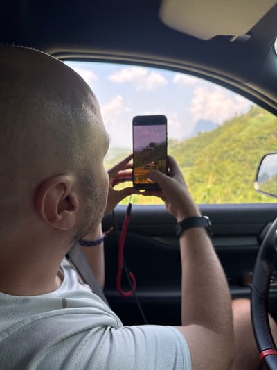 A man is seated in a car, holding a smartphone in his hand, taking a picture or video of the scenery outside the window. The exterior view is of green vegetation, possibly trees, under a sky with some clouds. The interior shows the car's steering wheel, part of the dashboard, and the side mirror. The man is wearing a white shirt and a seatbelt. A red cable is visible near his arm. The location could be Mậu Duệ Commune, Vietnam, based on external information, as this detail is not visually discernible from the image alone.