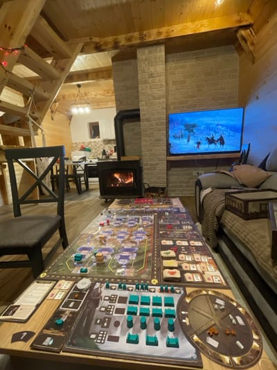 This image displays an interior view of a living space within a dwelling in Palež, Montenegro. The central feature is a large, multi-component board game, identified as "Barrage," fully arranged on a wooden coffee table. The game setup includes numerous teal, red, white, and brown tokens, cards, and miniature structures spread across the board's various sections. To the immediate left, a dark-stained wooden chair with a cushion is partially visible. To the right, a sofa draped with brown blankets and pillows holds a small, rectangular wooden tray. Directly behind the coffee table, a black wood-burning stove contains active flames and is positioned against a light-toned brick wall. A stack of cut firewood rests on the floor beside the stove. Above the stove, a flat-panel television is mounted, displaying a winter landscape with figures on horseback. The room features exposed wooden ceiling beams and paneling, with a wooden staircase ascending on the left, adorned with a string of red lights and a decorative hanging element. In the background, a kitchen area with white cabinetry, a counter, and a dining table with chairs is visible, illuminated by a multi-bulb chandelier. No individuals are present in the scene.
