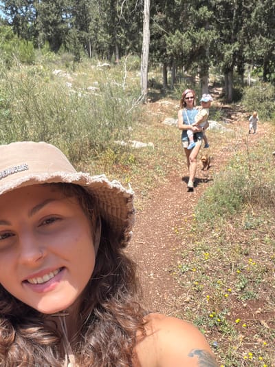 The image shows a smiling woman in a hat taking a selfie on a trail in what appears to be a natural area, possibly near the city of Zvulun Regional Council, Israel. In the background, another woman carries a child on the same path. The environment is characterized by trees, vegetation, and a dirt trail.