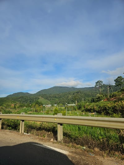 The image shows a roadside view in Klong Klanh, Vietnam. In the foreground, there is a gray asphalt road with a white line marking. A metal guardrail with concrete supports runs parallel to the road. Beyond the guardrail, there is tall green grass and dry brush. In the middle ground, a lush green hillside is covered with trees. Several buildings with light-colored roofs are scattered among the trees. The background features a large, mist-covered mountain range with a blue sky and scattered white clouds above. The scene appears to be a natural landscape with some signs of human habitation.