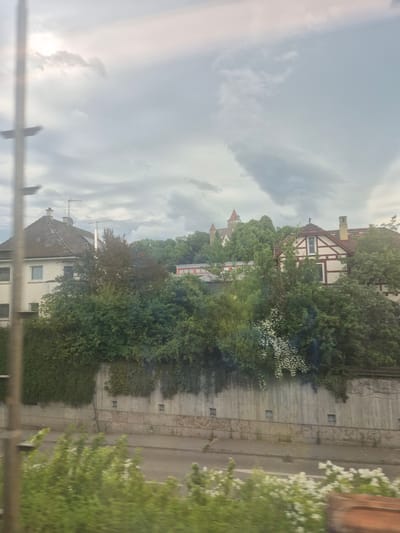 The image shows a view of Möckmühl, Germany, with buildings, trees, and a cloudy sky. A half-timbered house is visible near the top, and below it a castle turret. A road and a retaining wall appear on the right side. The overall environment suggests a mix of residential and natural elements within the city. Some railing is visible across the very bottom, obscuring the view.