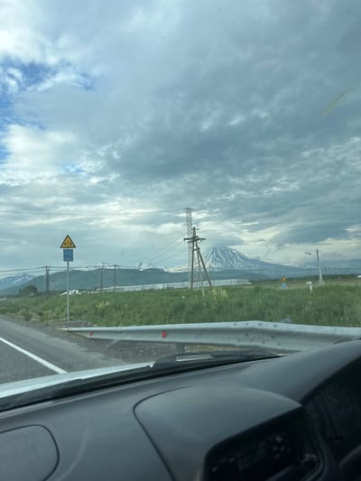 The image shows a view from inside a vehicle, looking out at a cloudy sky and a mountainous landscape. In the foreground, the dashboard of the car is visible. A road with a white line runs along the left side of the frame, with a metal guardrail to the right. There are power lines and utility poles scattered throughout the middle ground. A triangular yellow warning sign with an image of a person and a smaller blue sign below it is mounted on a pole to the left of the road. Further in the distance, snow-capped mountains are visible under a heavy cloud cover. There are no people or animals visible in the image. The scene appears to be in a rural or semi-rural area, likely on the outskirts of a settlement. The overall impression is of a somewhat overcast day in a natural setting with infrastructure elements. The presence of the warning sign and power lines suggest human activity in the vicinity. Given the context of Кеткино, Russia, this scene likely depicts a view of the surrounding natural terrain and infrastructure in that region.