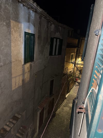 The image shows a narrow street view looking up in Castelnuovo di Porto, Italy, at night. Tall, old buildings flank the street, featuring weathered stucco walls, dark green shutters on windows, and tile rooftops. Electrical wires run along the walls. Overhead, a green metallic awning is visible. The scene is dimly lit, with some light emanating from the upper windows of the buildings, illuminating the cobblestone pavement.