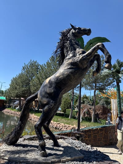 The image shows a statue of a rearing horse in Kenbulun, Kyrgyzstan. The statue is dark-colored, possibly bronze or a similar metal, and appears to be standing on a stone or rock-like base. Behind the statue, there is a park-like setting with trees, including palm trees. In the background, there is a body of water. Part of a building or structure is visible in the upper right corner, with someone standing on top of it. The sky is clear and blue.