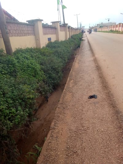 The image depicts a street scene in Tangu, Nigeria, likely during the daytime under an overcast sky. On the left, a low wall made of concrete and brick runs along the edge of the property, topped with barbed wire for security. Adjacent to the wall, dense green foliage spills over and into a drainage ditch. The ditch itself is partially filled with mud and some debris, including a black plastic bag lying on the gravel shoulder of the road.

The road, paved with a reddish-brown material, stretches into the distance and appears to be moderately busy. Several motorcycles are visible on the road, with riders on them. In the background, there are buildings with red-tiled roofs and more walls lining the street, suggesting an urban or semi-urban environment. Utility poles and streetlights are also present. The overall atmosphere is utilitarian, with no overt signs of celebration or distress, but the presence of the barbed wire and the rough state of the drainage ditch might imply a less developed or well-maintained area. There is no visible text in the image.