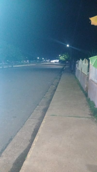 The image depicts a street scene in El Samán, Venezuela at night. Streetlights illuminate the road, which is adjacent to a sidewalk. Some low buildings or structures are visible alongside the sidewalk. The sky is dark. No people or vehicles are visible.