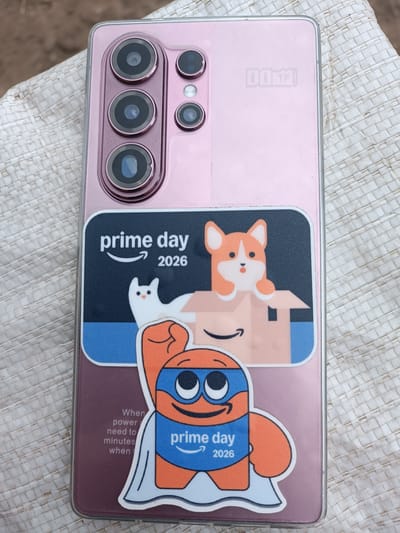 The image shows the back of a pink smartphone with a transparent case. The phone has a prominent camera module with three circular lenses arranged vertically, and two smaller circular lenses and a flash to the right of the main module. A faint logo, possibly "DDM," is visible on the back of the phone.
A large sticker covers the lower half of the phone's back. The sticker features several graphics. The top part of the sticker has the text "prime day 2026" in white lettering against a dark blue background, with the Amazon smile logo. Below this text are cartoon illustrations of a white cat and a corgi dog peeking out of a cardboard box. The bottom part of the sticker shows a cartoon character wearing a blue mask and a blue cape with the Amazon logo and the text "prime day 2026." To the left of this character, small white text reads "When power need to minutes when".
The phone is placed on a woven, light-colored surface, possibly a placemat or a fabric mat. The background is slightly blurred, with hints of brown and green, suggesting an outdoor or natural setting. The overall impression is of a product photograph, likely taken to showcase the phone and its accessories or decorations. There is no visible indication of the city of Leușeni, Moldova, or any specific urban environment.