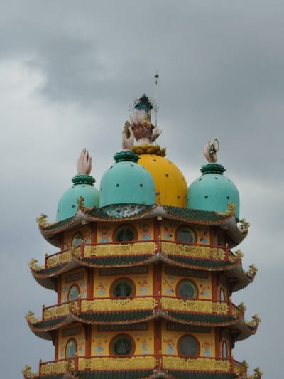 The image shows the top of a multi-tiered pagoda in Thiện Chí, Vietnam, under a cloudy sky. The pagoda features a yellow exterior with red and gold detailing. Each tier has small, round windows and green tiled roofs. The top of the pagoda has three turquoise domes and one yellow dome. Large hands, seemingly part of statues, are positioned on top of each dome. One statue is holding a lantern-like object.