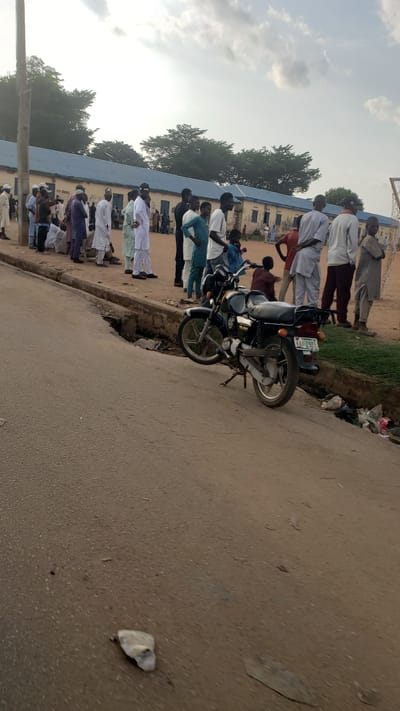 The image shows a street scene in Kawo, Nigeria. A group of people is lined up along a building with a blue roof. The people are wearing a variety of clothing. A motorcycle is parked on the edge of the road, near a curb. The road surface appears to be paved, and there is litter present. The sky is visible, with clouds. The image is rotated.