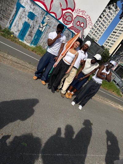 Six young individuals are posing diagonally across a paved outdoor area under bright sunlight in Charenton-le-Pont, France. The group consists of four males and two females, all appearing to be of Black ethnicity, dressed in casual streetwear. Their attire predominantly features white tops, baggy jeans or shorts, and accessories such as hats, sunglasses, and chains. Many are smiling or striking poses for the camera.
To their left, a white wall is extensively covered in red and grey graffiti, depicting a stylized skull or face and various abstract patterns. Words like "VIEJO" appear to be part of the graffiti. Behind this wall, tall, modern multi-story residential buildings rise against a clear blue sky. To the right of the subjects, there's a paved road or path with a curb, patches of grass, and a metal fence.
One woman in the center of the group is holding an orange and white flag. Strong, elongated shadows are cast on the pavement, indicating a sunny day, likely morning or late afternoon. The overall atmosphere is cheerful and relaxed, capturing a moment of social interaction in an urban setting.