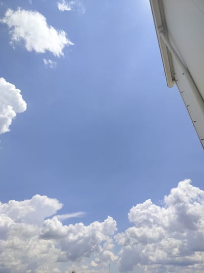 The image depicts a bright blue sky with scattered white cumulus clouds. In the upper right corner, a portion of a white building or structure with a gutter system is visible, angled downwards towards the sky. The lower part of the image is filled with more dense clouds, some appearing dark and heavy, suggesting potential for rain, while others are bright and fluffy. At the very bottom edge, what appear to be the tops of industrial structures or antennas can be faintly seen emerging from the clouds. The scene is a clear, daytime shot focusing on the sky and a partial view of an urban or industrial edge. There are no people or discernible actions visible.