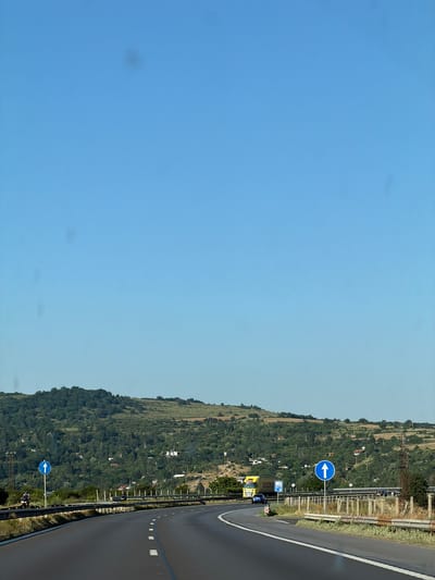 This media file depicts a highway scene in Eleshnitsa, Bulgaria. The image is oriented vertically, showing a paved road curving to the right, bordered by a metal guardrail. Beyond the guardrail, a densely vegetated hillside rises, dotted with scattered houses. The sky is clear and a light blue, suggesting daytime with fair weather.
Notable objects in the scene include traffic signs. A circular blue sign with a white arrow pointing to the left is visible on a pole beside the road. Further down the road, a yellow truck with the word "DISOPO" printed on its side is parked, and a person in a blue shirt is standing nearby. Another blue sign with a white arrow is visible further along the road. Power lines and a transmission tower are also present on the hillside.
The overall impression is one of a scenic, rural highway on a clear day. There are no strong indicators of a specific event or activity other than general traffic and the presence of a parked truck. The time of day is likely afternoon, given the lighting. The location cues are the specific town name provided and the visual characteristics of the Bulgarian landscape, which include a hilly terrain covered in lush greenery and a typical European-style highway with its accompanying infrastructure.