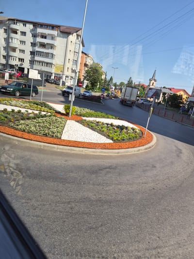 This is a medium shot, taken from a slight upward angle, of a street scene in Fălticeni, Romania. The foreground is dominated by a well-maintained roundabout with colorful flower beds and decorative gravel. The roundabout is bordered by a curb and paved road. In the middle ground, traffic is visible. A green station wagon is turning into the roundabout, and further into the scene, a white van and a white sedan are driving on the road. A large white truck is also moving away from the roundabout. On the left side of the street, a modern apartment building with multiple balconies and windows stands tall. Across the street, a church with a tall steeple and red-tiled roofs is visible, along with other buildings. The sky is clear and blue, indicating a sunny day. The overall scene depicts a typical urban environment with active traffic and residential and commercial buildings.