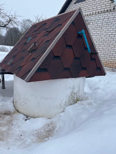 The image displays a well in a snow-covered outdoor environment, located in Карабоўскі сельскі Савет, Belarus. The well consists of a cylindrical, white-painted concrete base, partially obscured by accumulated snow. The lower section of the base shows areas of discoloration, including greenish stains and dirt, particularly on the right side. Surmounting the base is a pyramidal, four-sided roof covered with reddish-brown and dark brown hexagonal asphalt shingles, finished with lighter brown trim along its edges. On the visible left slope of the roof, three metallic latches and a wooden handle secured with two visible bolts are present. On the visible right slope, a blue, L-shaped pipe or handle protrudes horizontally. The ground around the well is blanketed in white snow, with patches of disturbed, dirty snow and exposed brown earth visible directly surrounding the well's base on the left side. In the background to the left, bare deciduous trees are visible against an overcast, light grey sky, indicating a winter setting. To the right, a section of a light-colored brick building with white mortar lines is partially visible, featuring a dark roofline. No individuals or animals are present in the scene.